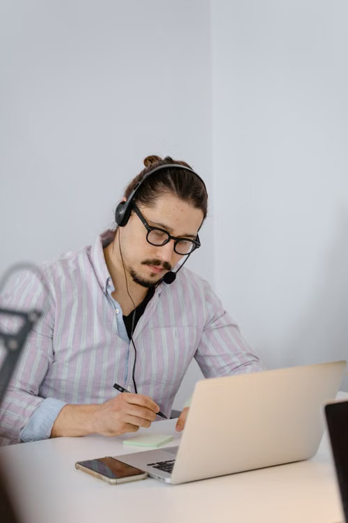 Professional in a headset taking notes beside a laptop during a certification course